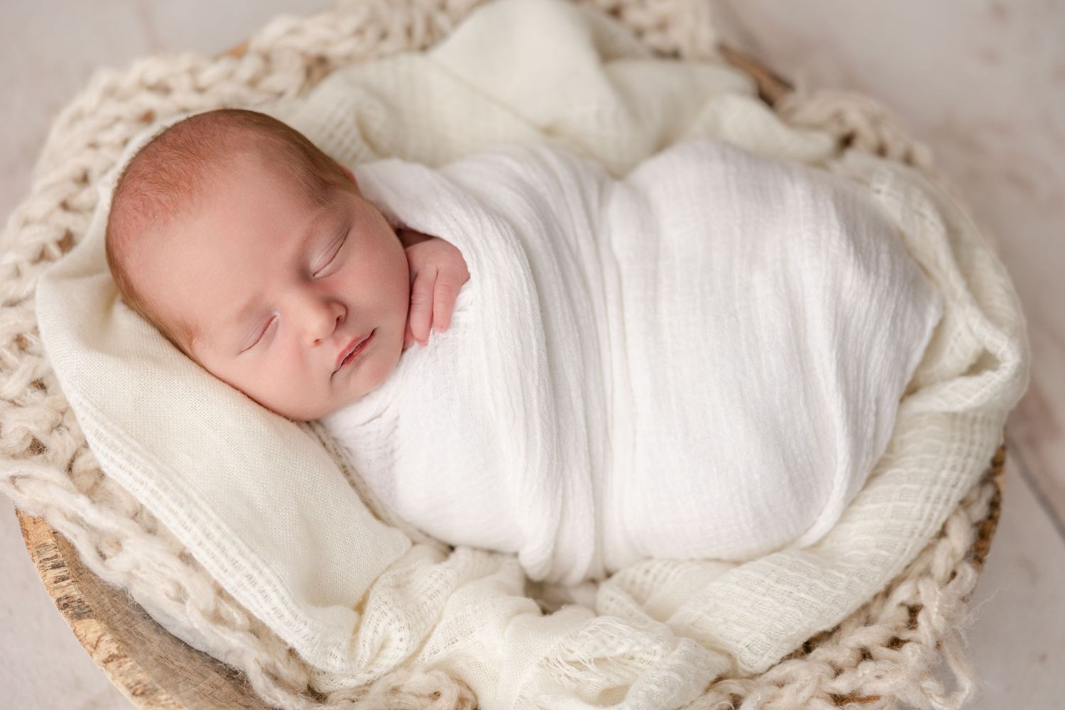 Newborn baby wrapped in white swaddle sleeps peacefully in vintage lace basket.