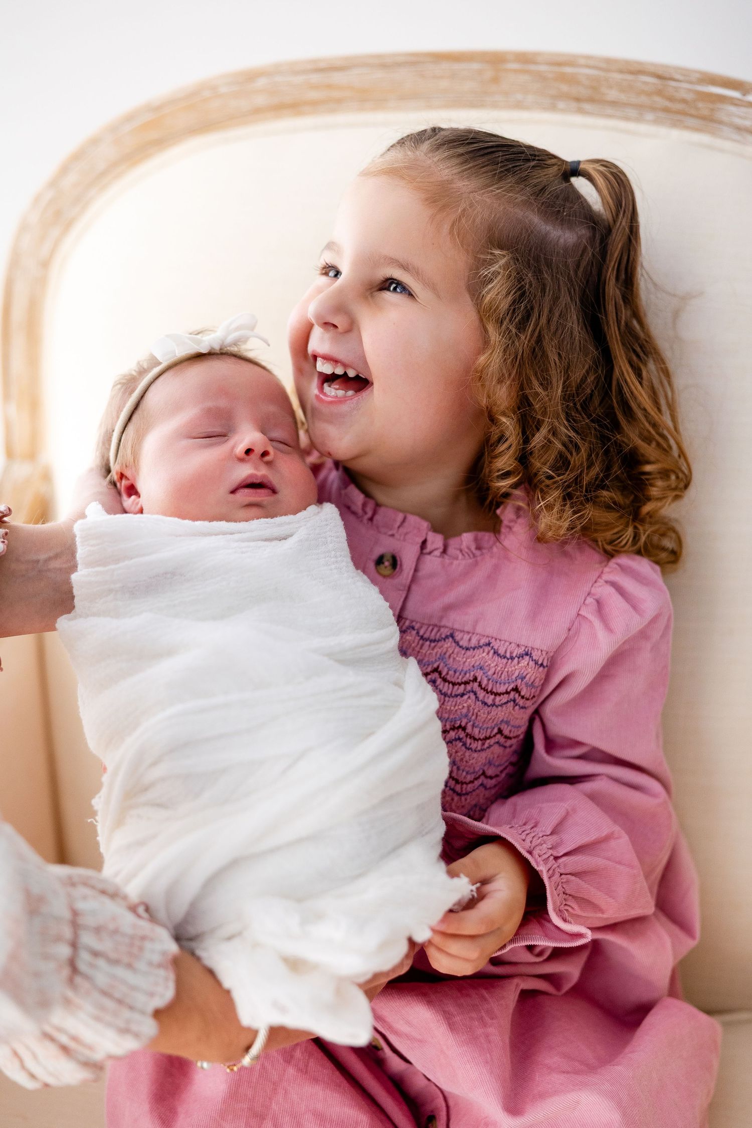 Two children share a tender moment on white bedding, with one wearing a pink dress and showing joyful expressions.