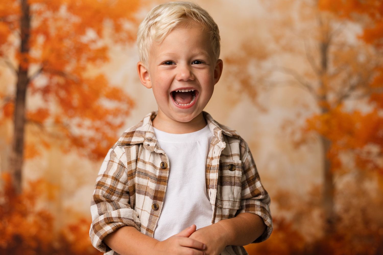 A joyful blonde boy laughs with arms crossed, wearing a plaid shirt against a vibrant autumn foliage backdrop.