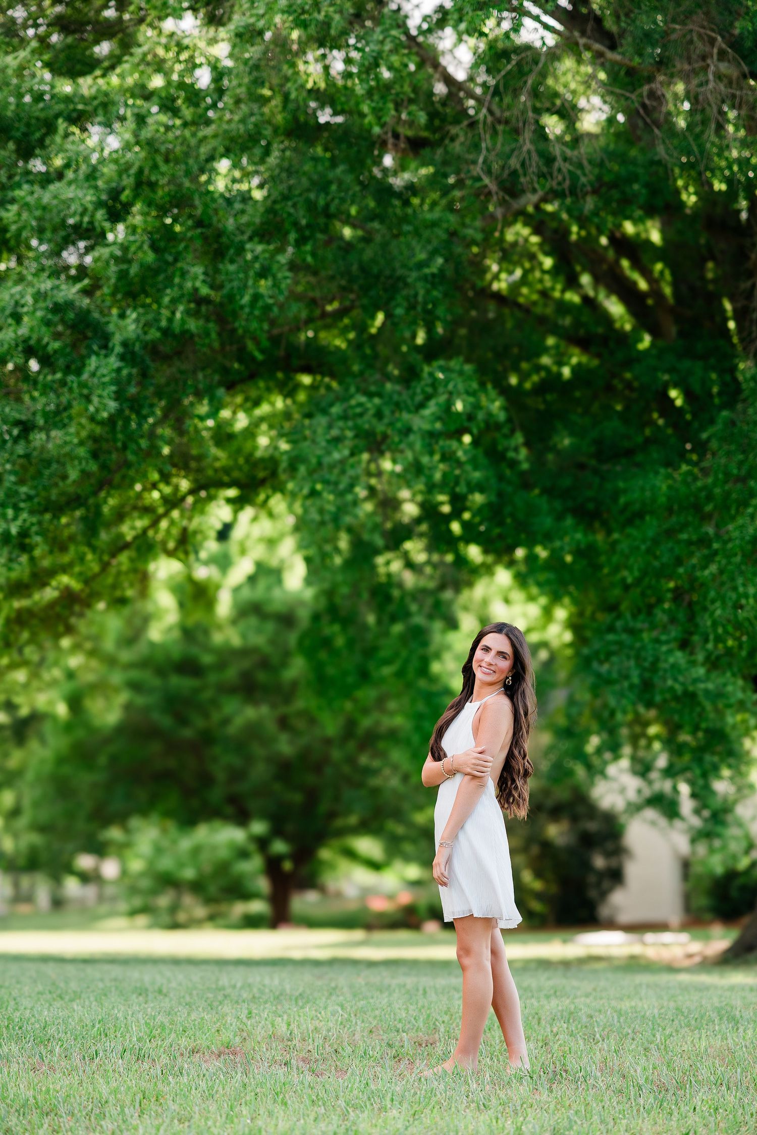 Young woman in a white sundress standing barefoot on green grass beneath large shady trees in a park.