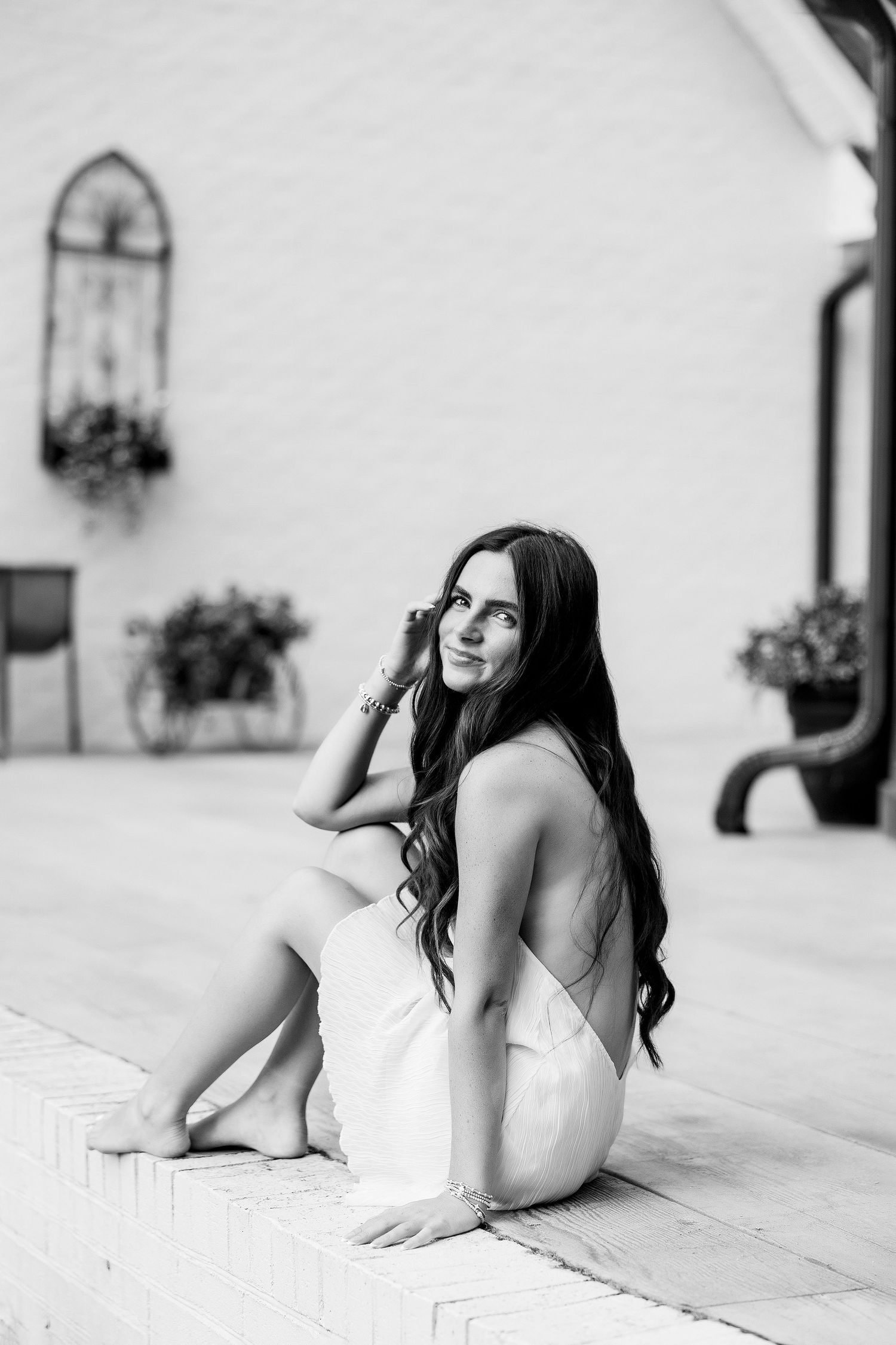 Black and white photo of a young woman with long dark hair sitting gracefully on a patio in a white dress.