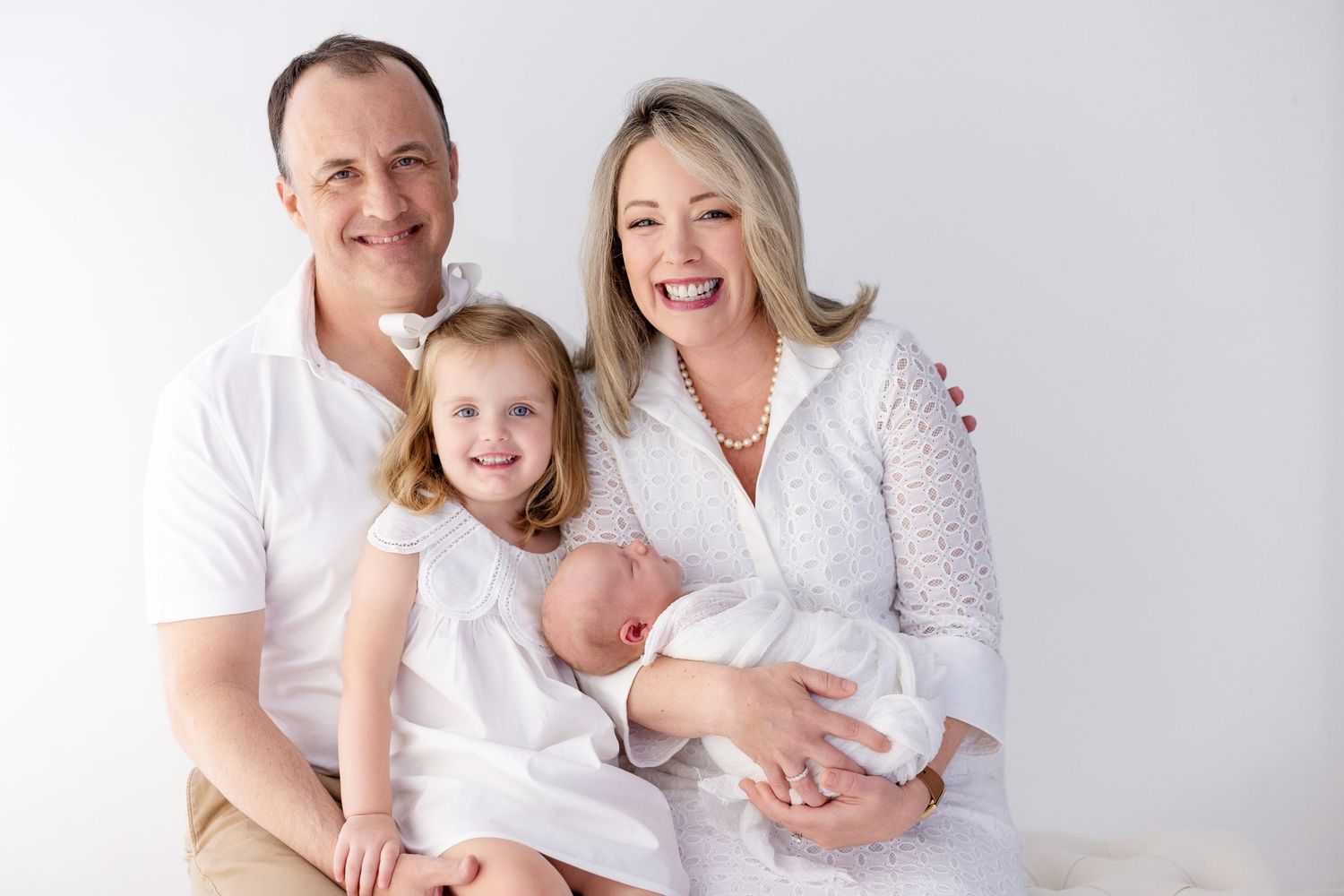 Happy family of four in white outfits posing together, parents smiling with young daughter and newborn baby.