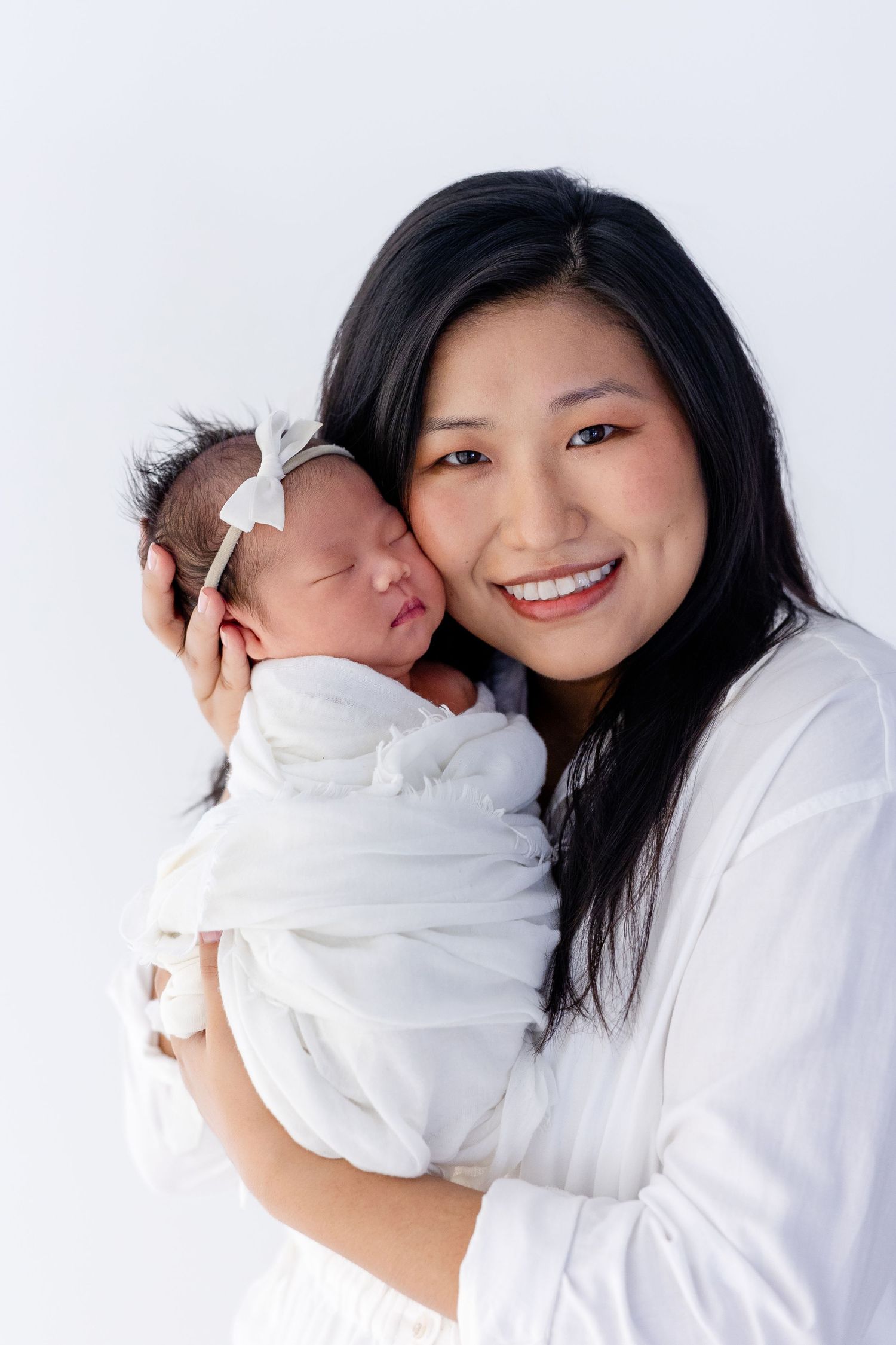Smiling Asian mother in white holding sleeping newborn baby girl with white bow headband against white background.