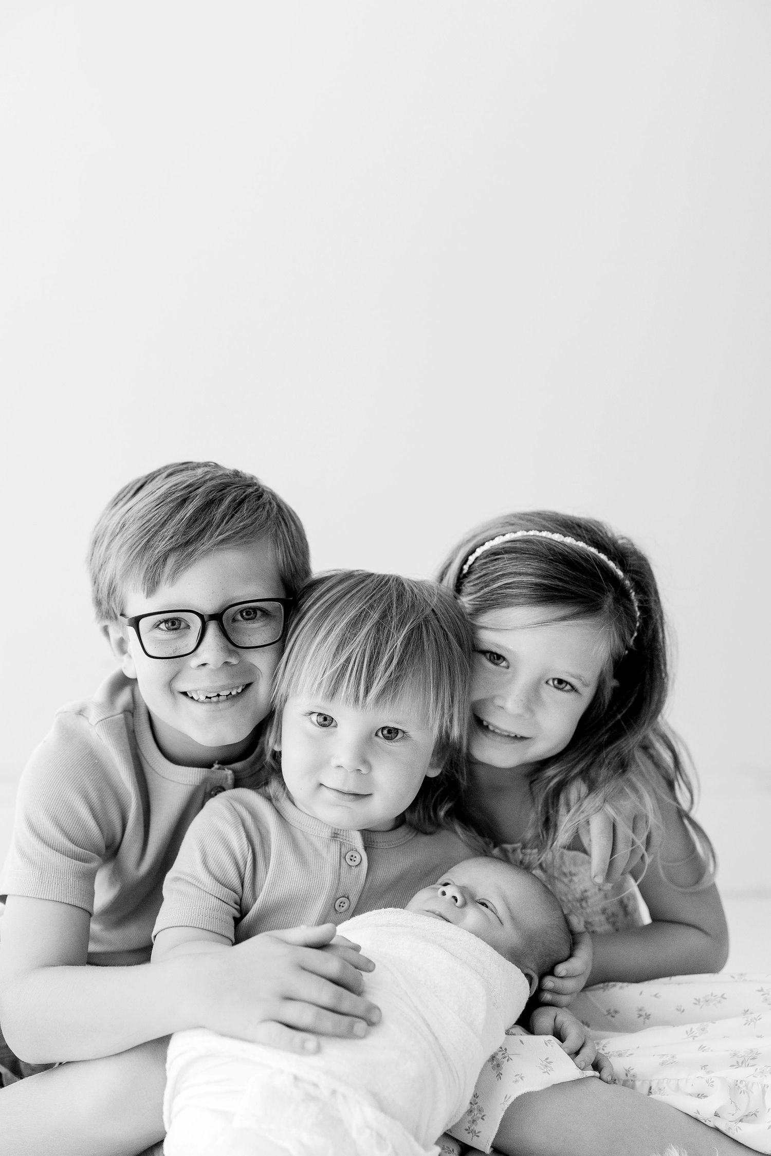 Black and white photo of four siblings, including a newborn baby, posing together for a family portrait.