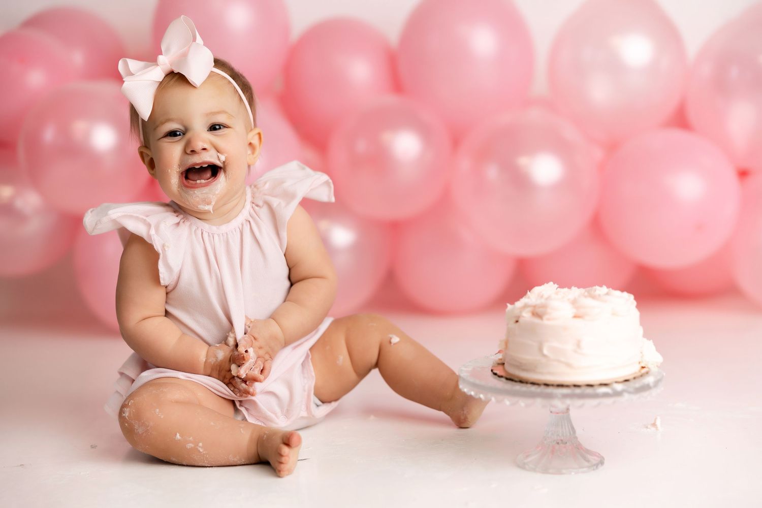 Baby in white outfit celebrates first birthday with pink balloons and smash cake in a sweet photoshoot setup.