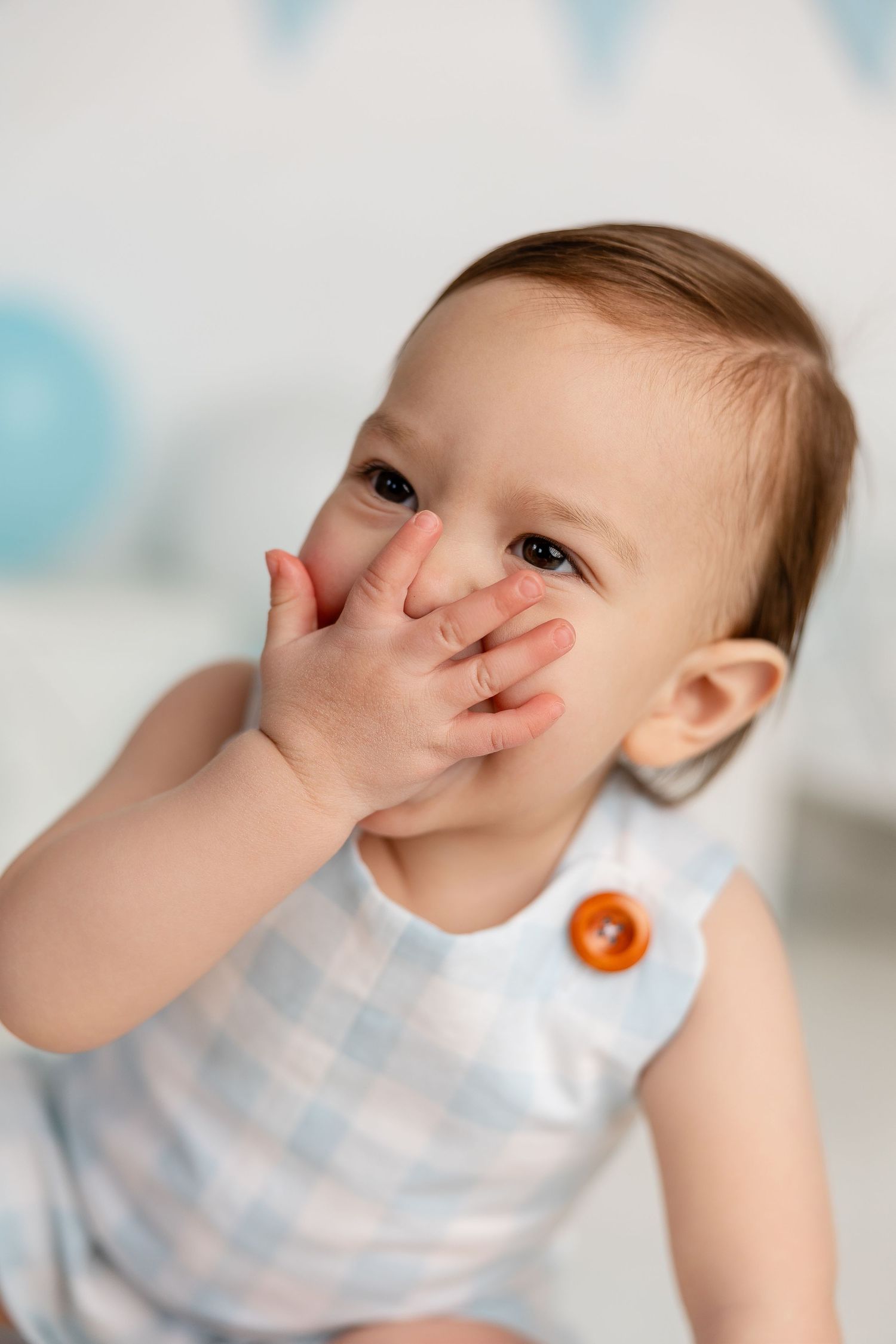 Baby in blue gingham romper playfully covers face with hands against white backdrop with blue decor.