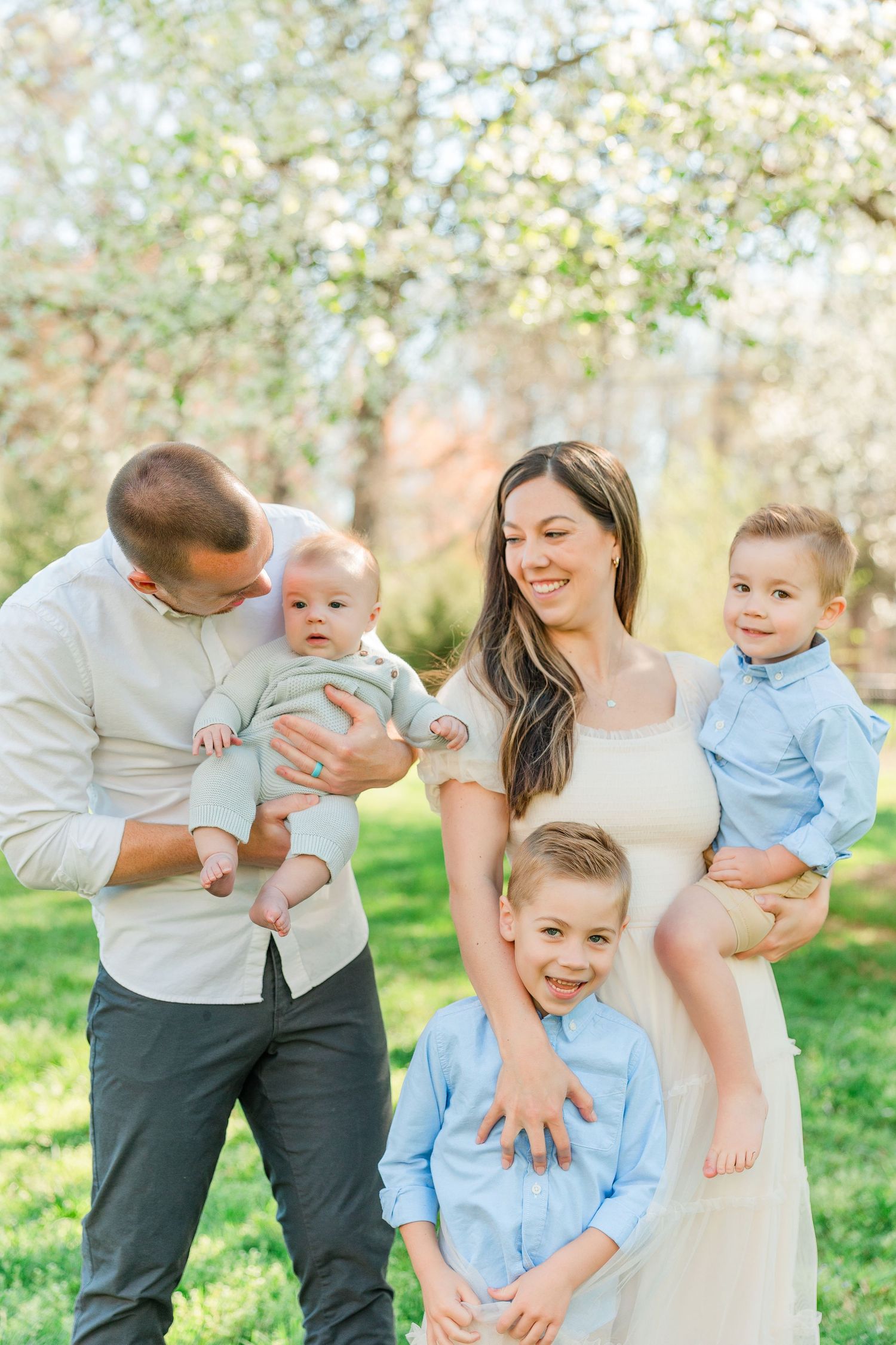 A family of five poses together outdoors during springtime with blooming trees in the background.
