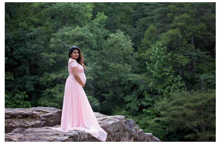 A flowing pink dress cascades over natural rock formations in a dreamy forest maternity photoshoot.