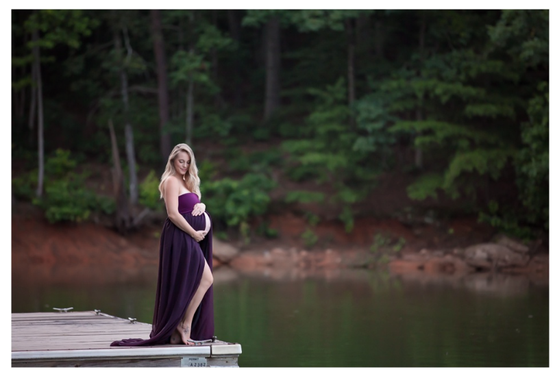 A pregnant woman in a flowing purple dress stands on a wooden dock by a serene lake surrounded by forest.