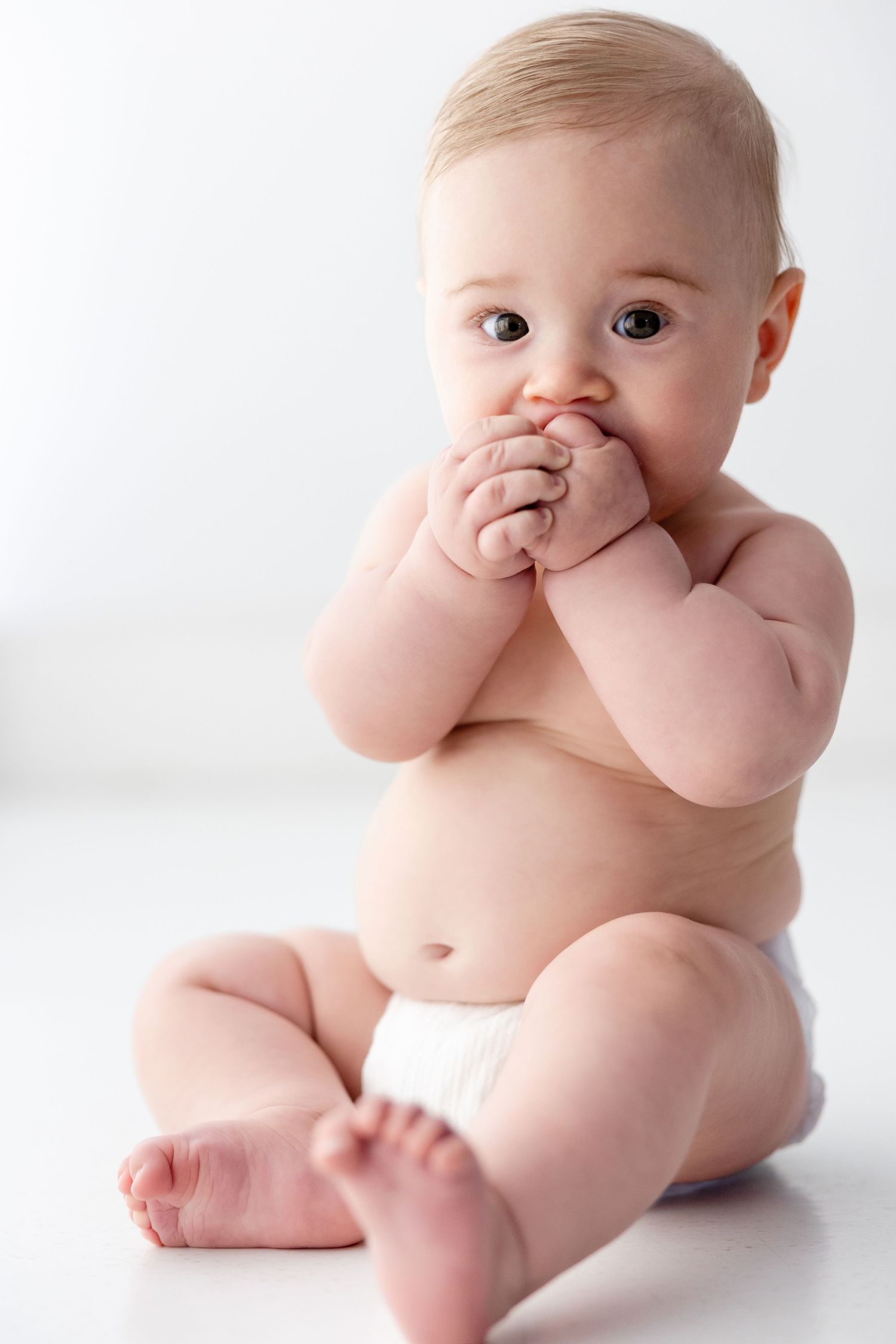 Baby sitting on white surface showing playful expressions while touching face.