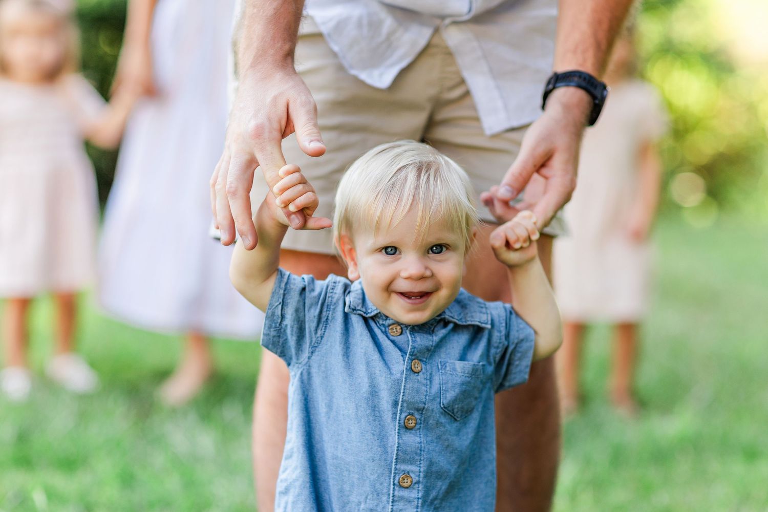 Baby in denim overalls taking first steps during outdoor family photo session.