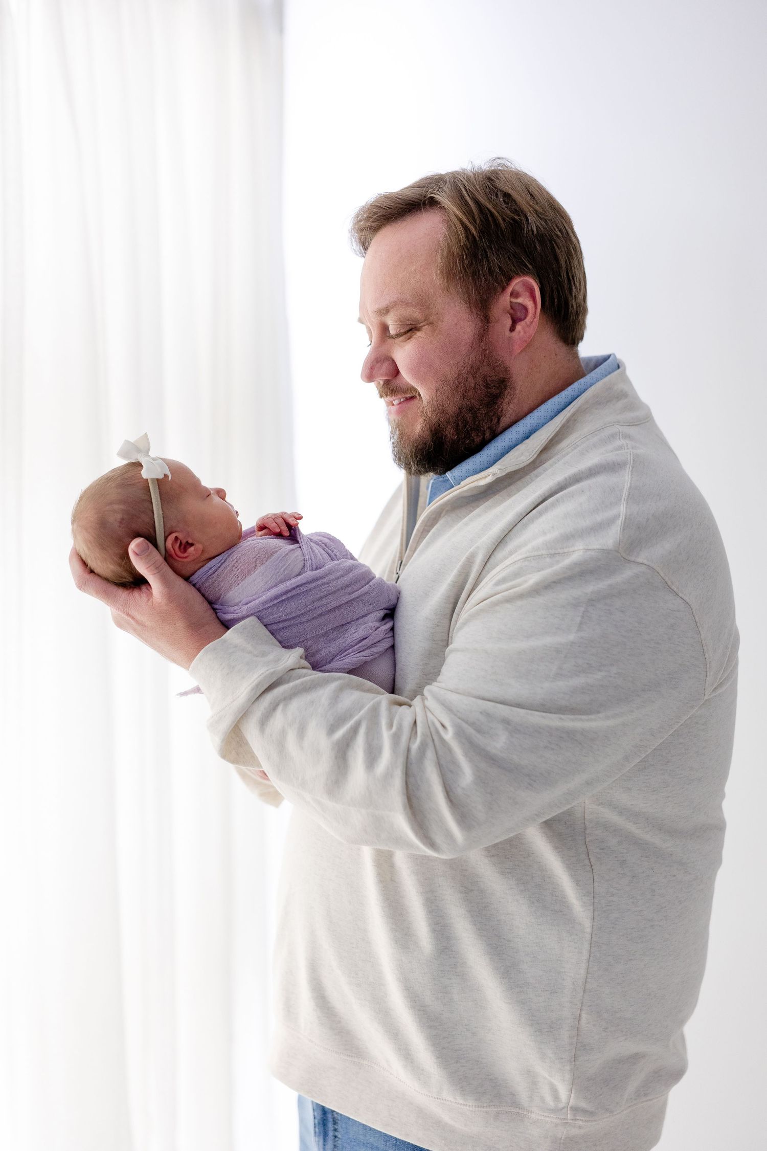 A bearded man in a white sweater tenderly holds a newborn baby wrapped in purple against a bright window backdrop.