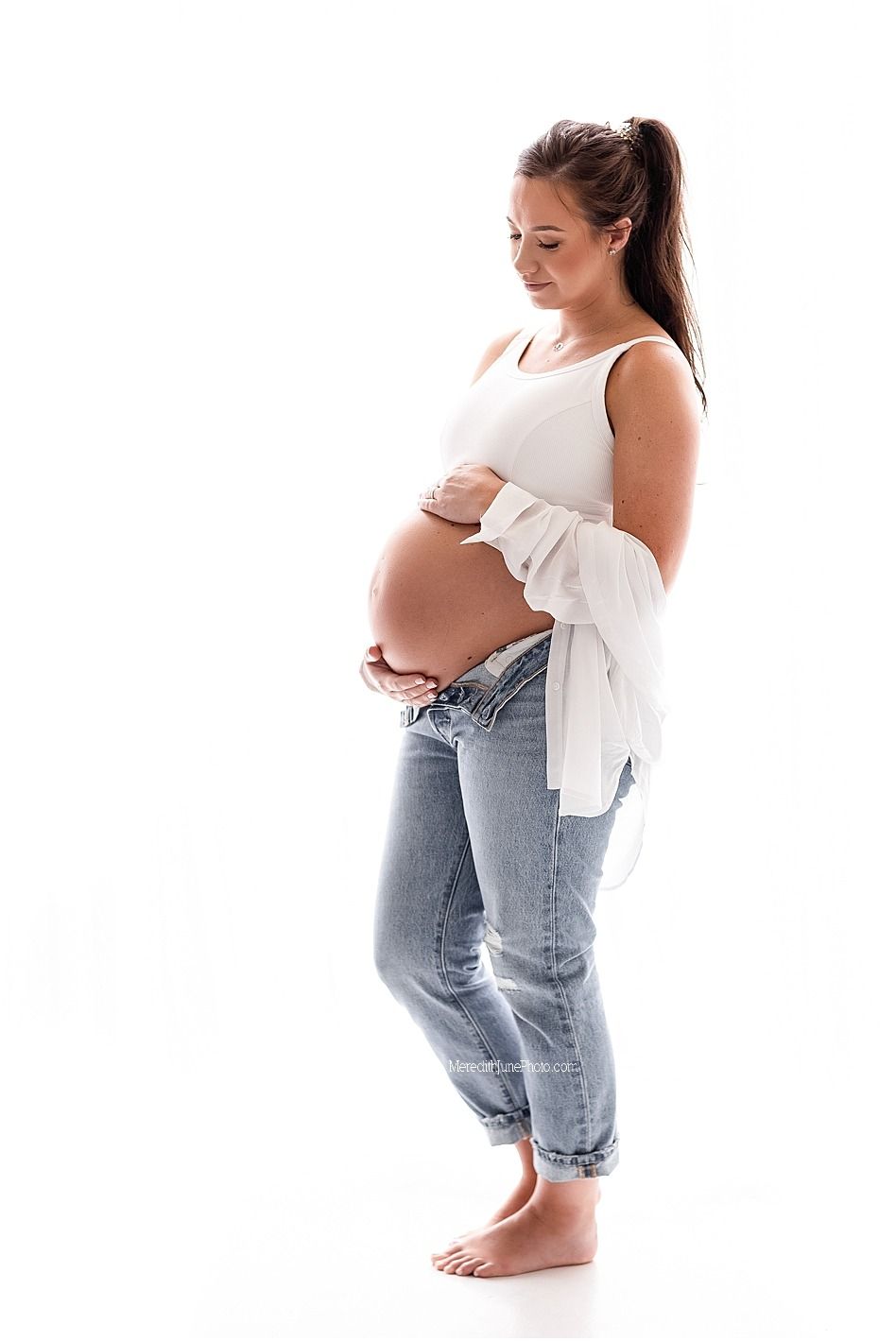 Expectant mother in white tank top and jeans stands barefoot against white background showing her pregnant belly.