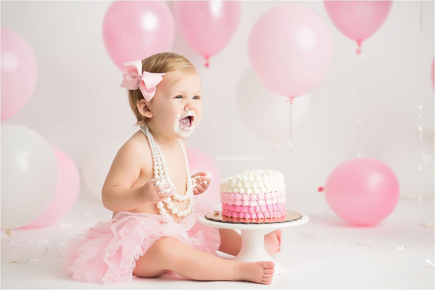 First birthday cake smash setup with pink balloons, tulle skirt and pearls against white backdrop.