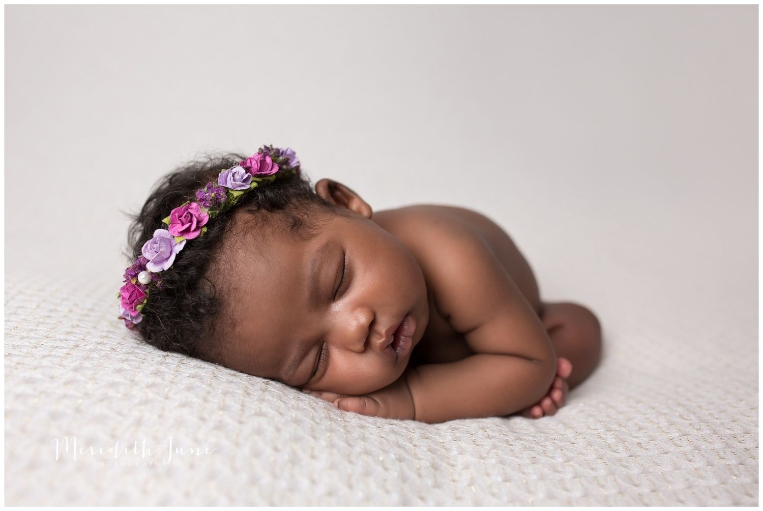 A sleeping newborn baby wearing a pink floral headband rests peacefully on a white backdrop.