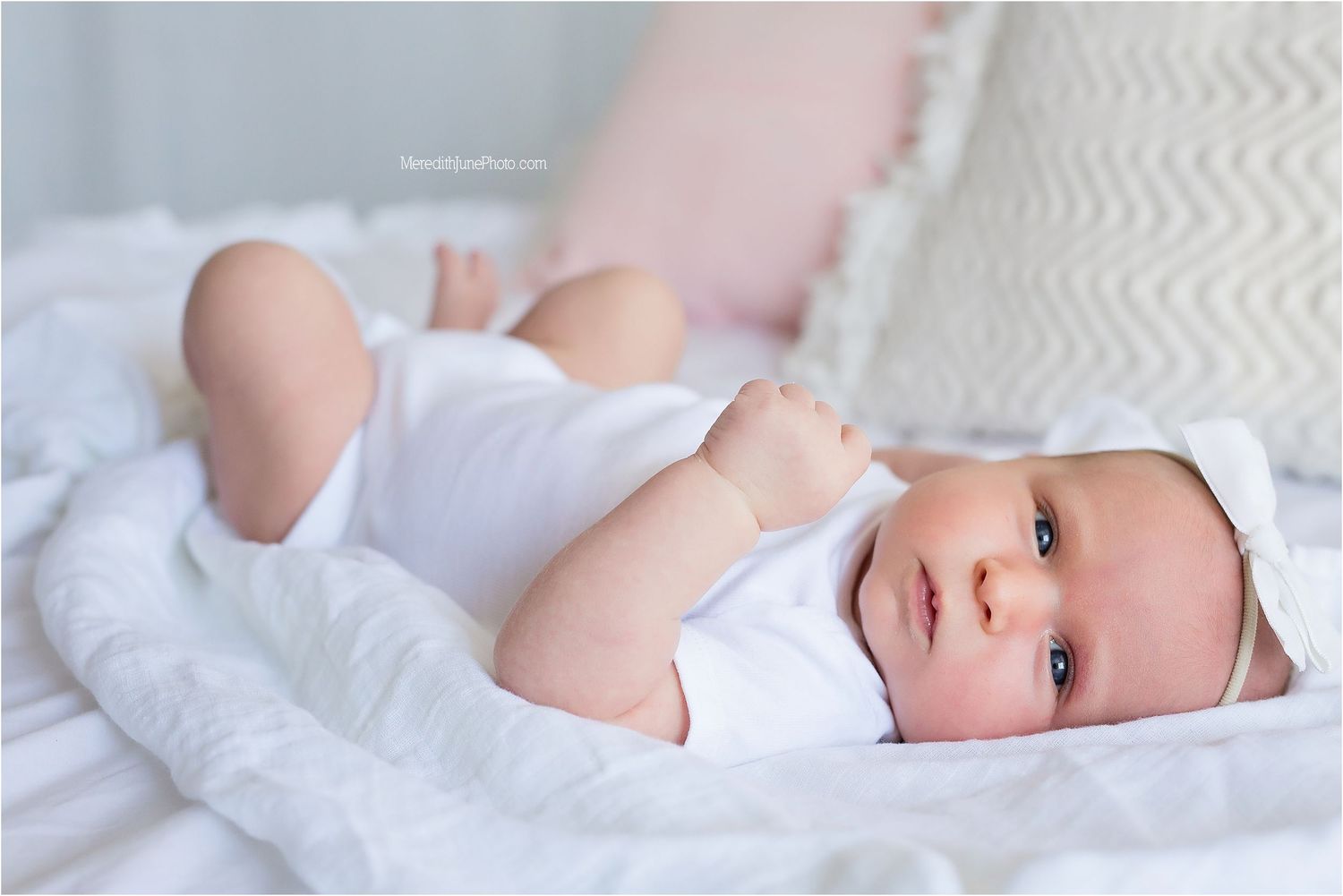 A newborn baby lies peacefully on white bedding while looking towards the camera with bright eyes.