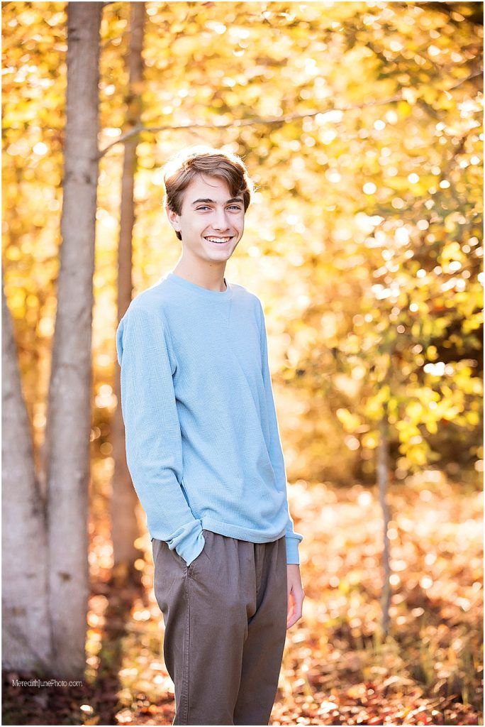 A person in a light blue sweater stands in a wooded area with golden autumn foliage in the background.