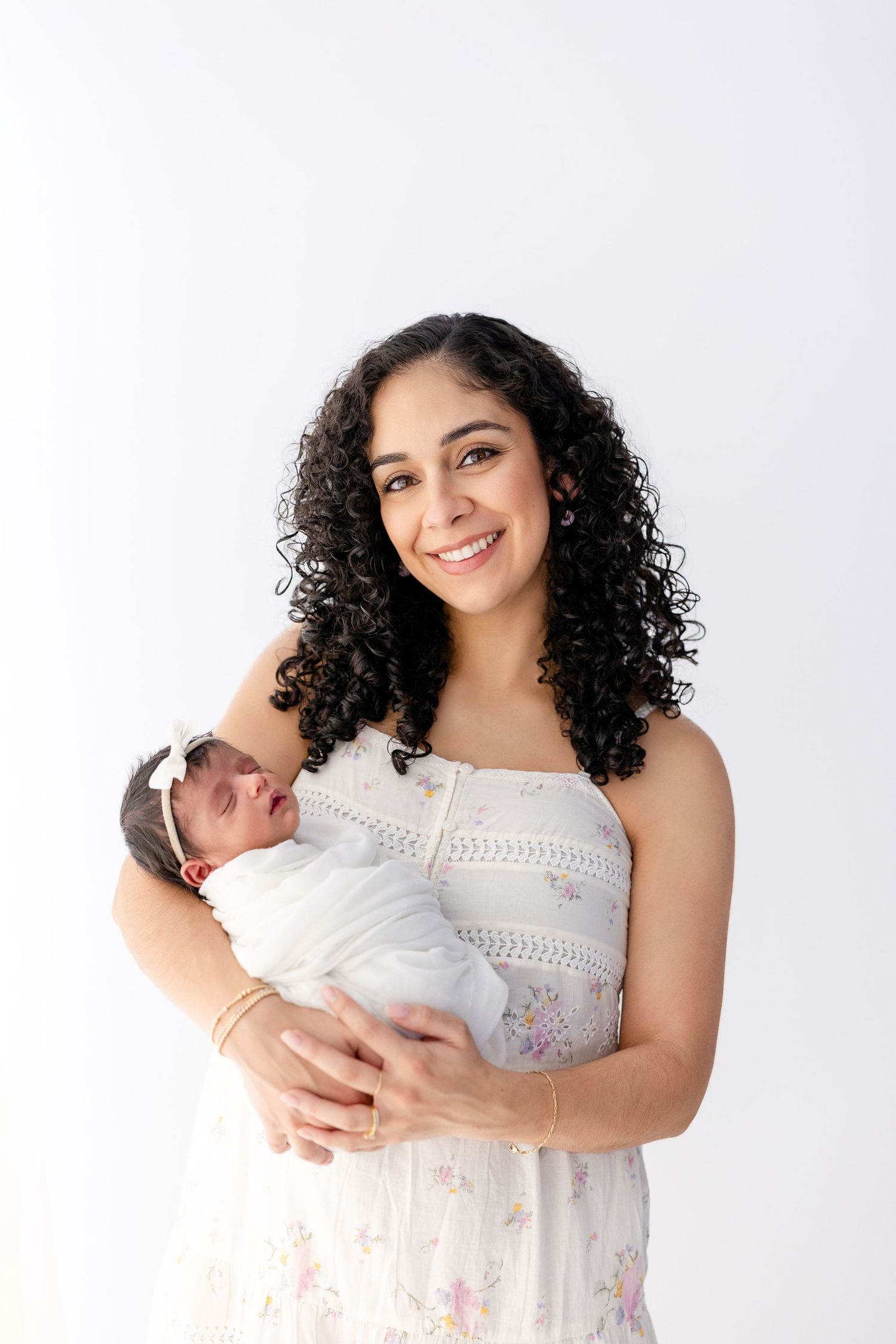 Mother in white lace dress holds newborn baby in series of studio portraits against white background.