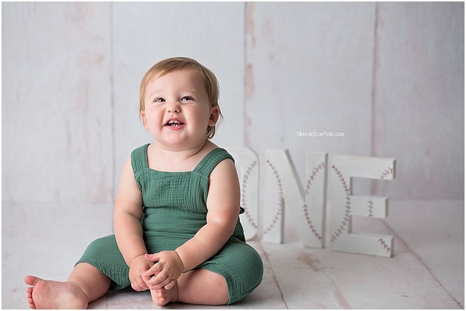 Baby in green romper sits and smiles on white backdrop with decorative elements.