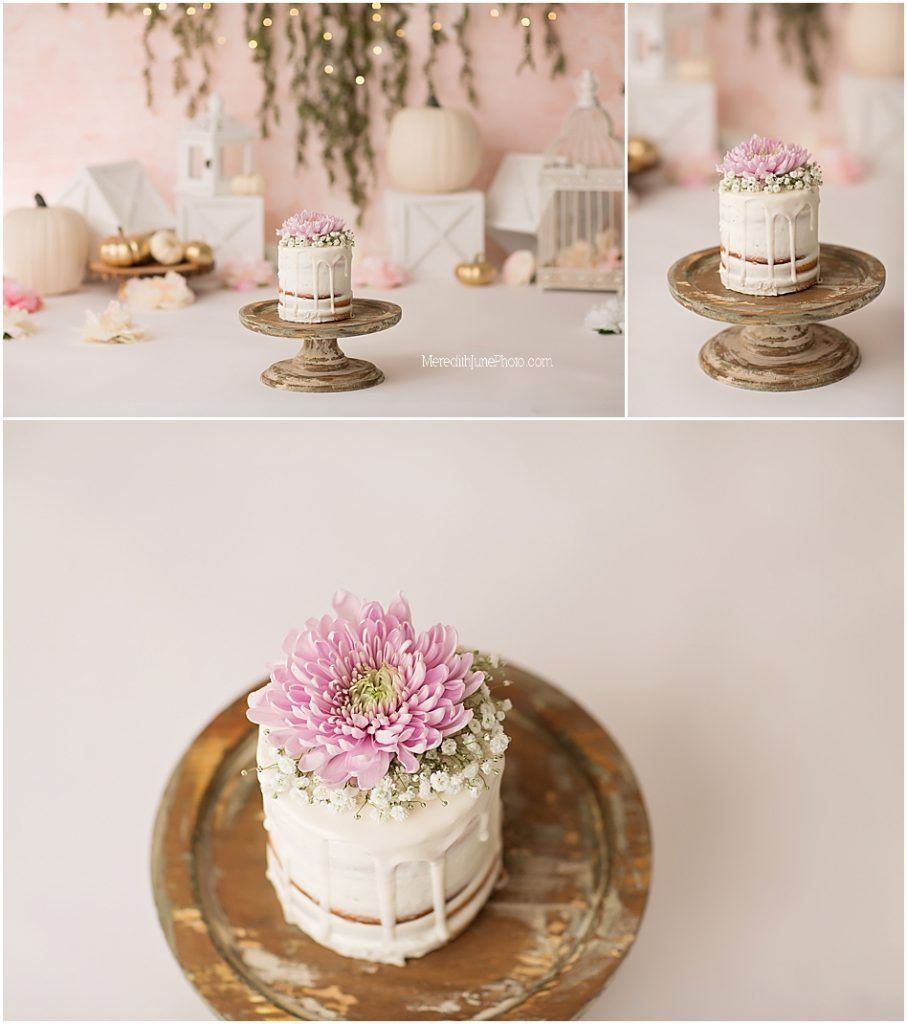 A rustic wooden cake stand displays a delicate white cake decorated with pink flowers and greenery in soft lighting.