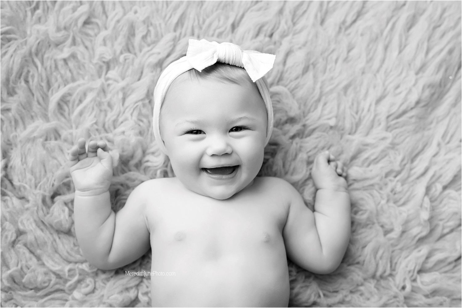 Black and white portrait of a smiling baby wearing a white bow while lying on a fluffy blanket.