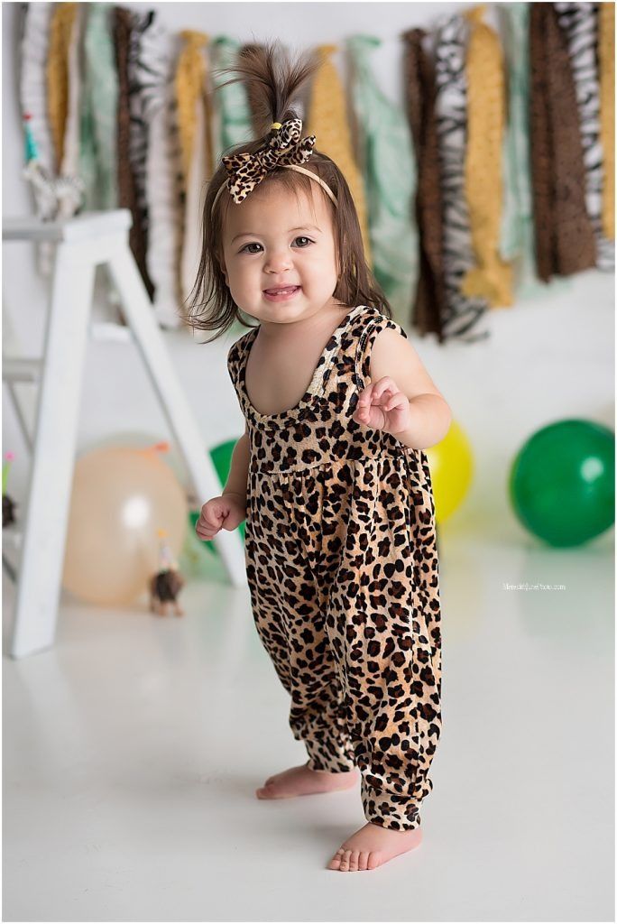 A small child wearing a leopard print romper poses playfully against a backdrop with colorful balloons.