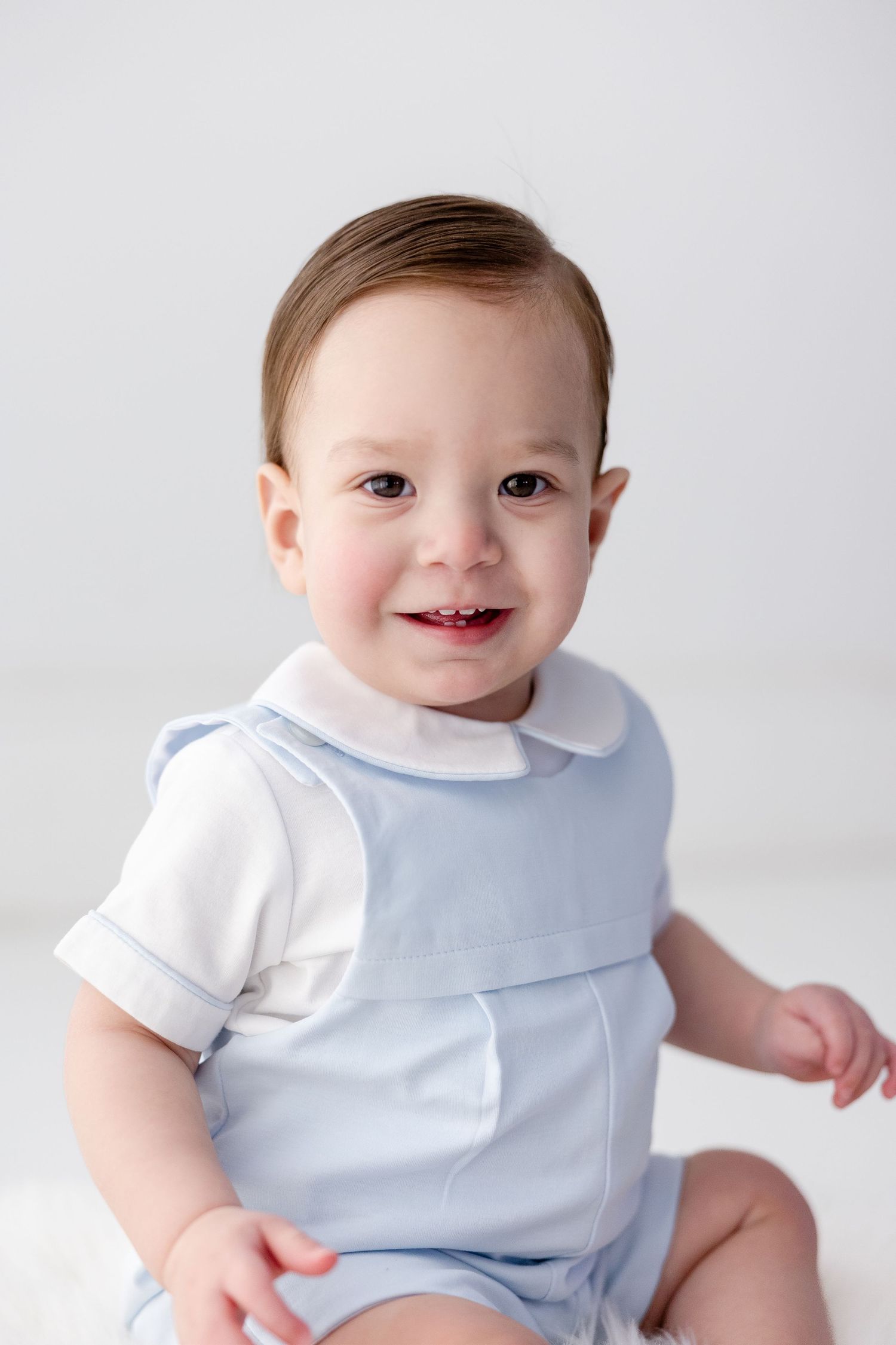 Baby wearing light blue bib and white shirt sits against white background in studio portrait series.