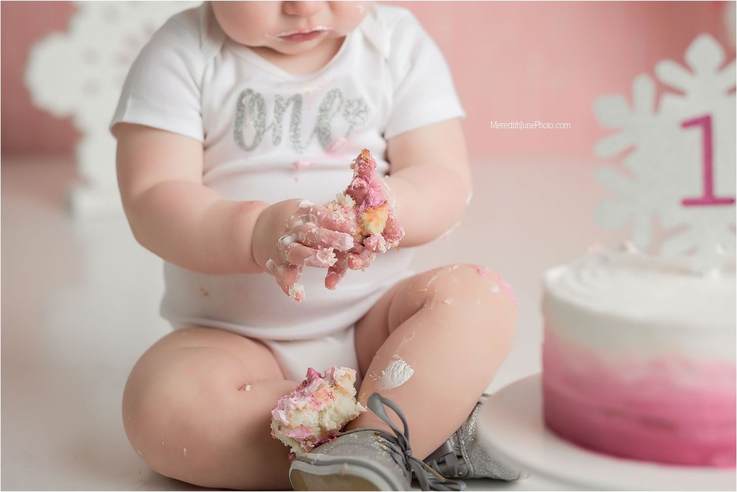 Small child in white 'one' shirt plays with pink frosting from birthday cake on white background.