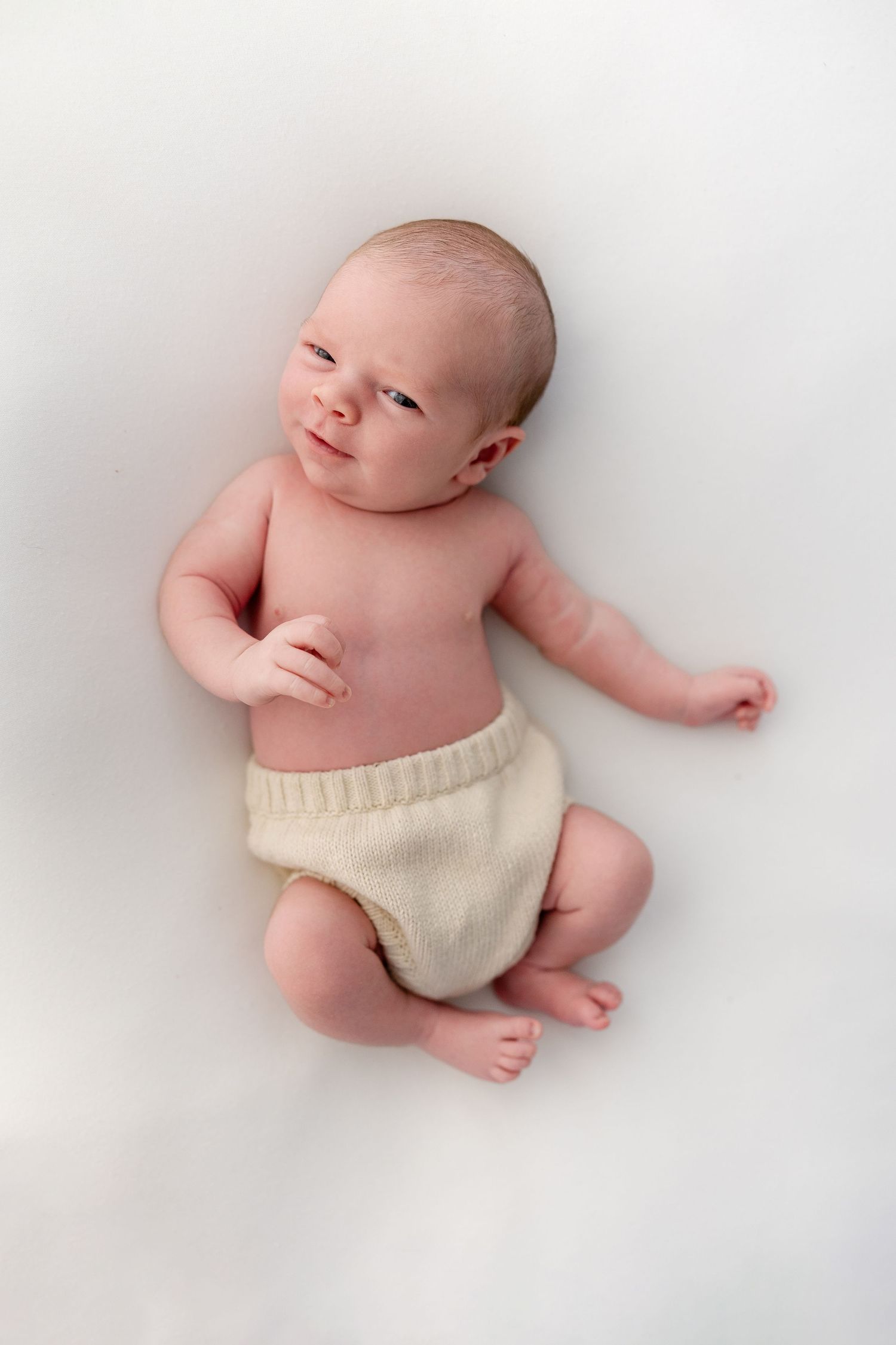 Newborn baby lying on white background wearing cream knit shorts, smiling softly with eyes open.