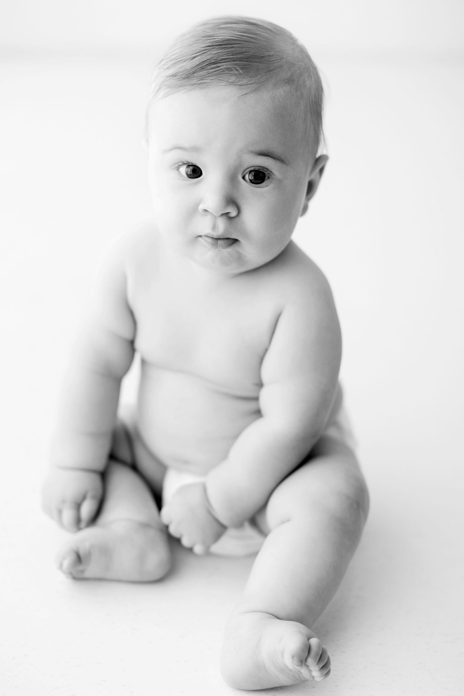 Black and white studio portrait of a baby sitting against white background in natural lighting.