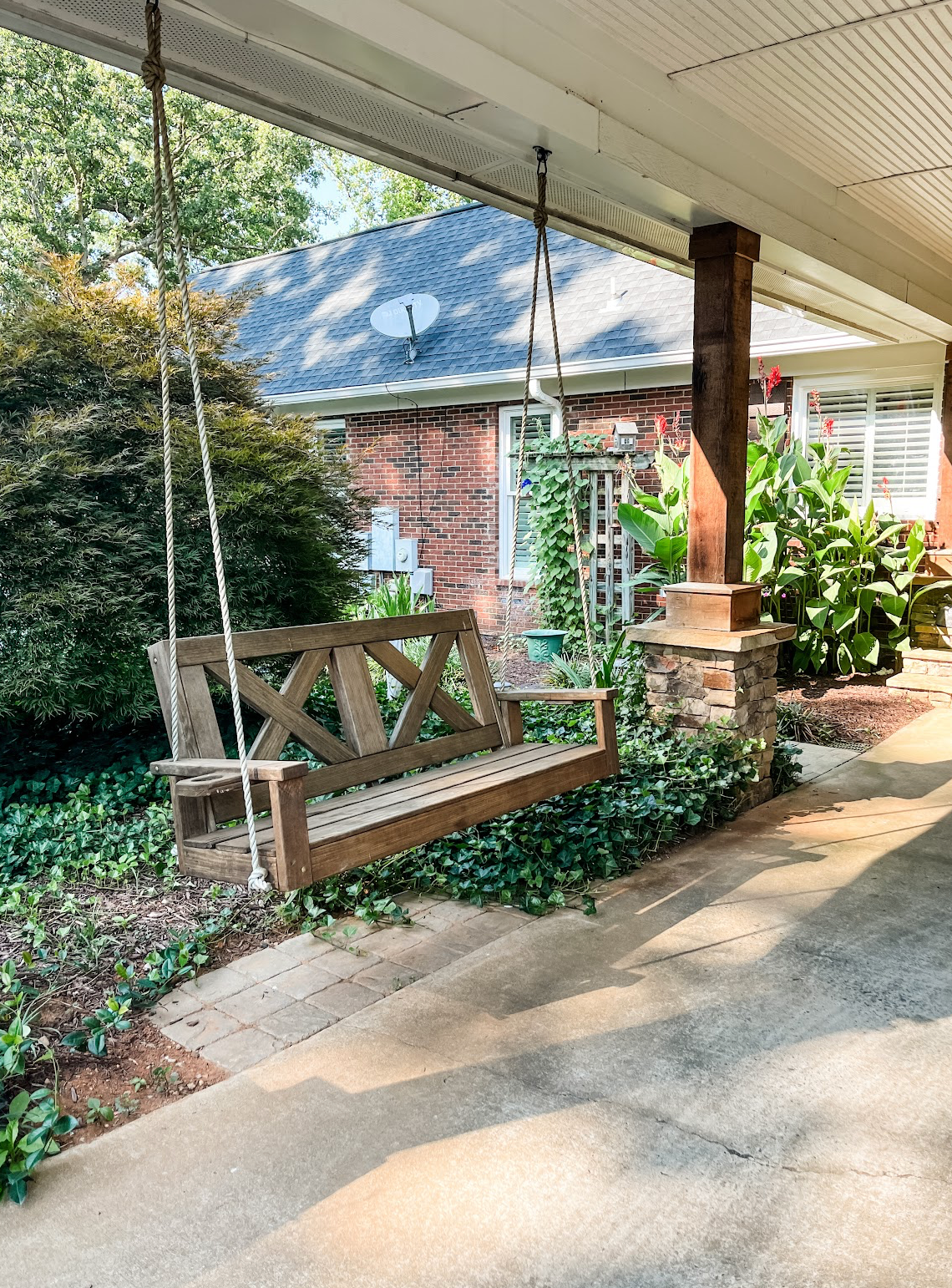 A rustic wooden porch swing hangs from a covered entryway of a brick house with lush garden landscaping.