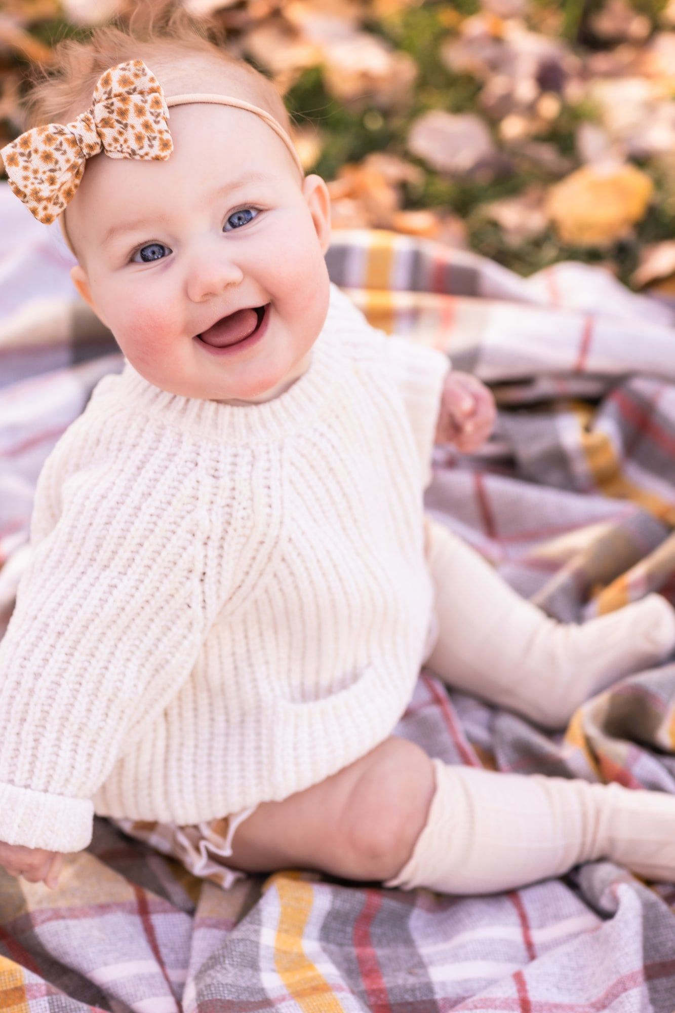 Baby in white sweater with bow headband sitting on plaid blanket among fall leaves.