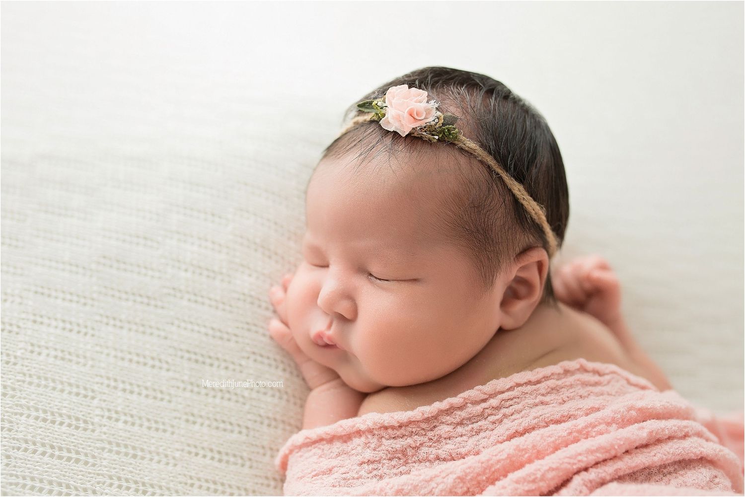 Sleeping newborn baby wrapped in soft pink blanket with delicate floral headband resting on white textured fabric.