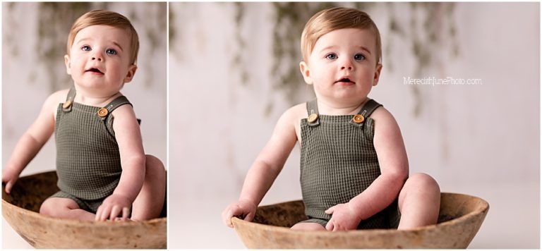 A small child wearing an olive green romper sits in a wooden bowl against a light background.