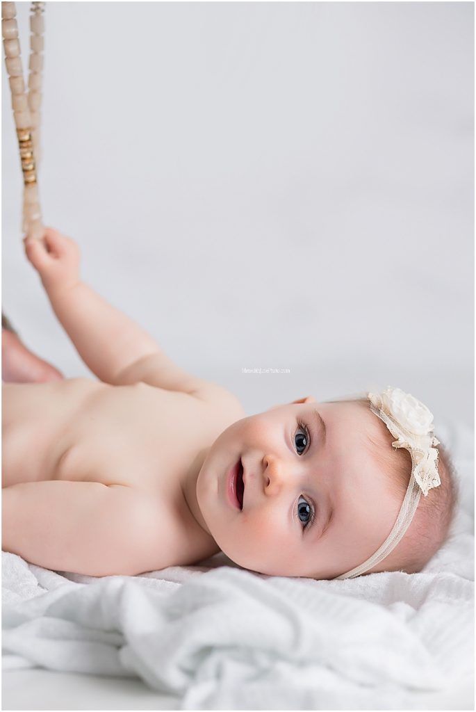 A baby wearing a delicate cream headband lies on soft white bedding in a series of gentle poses.