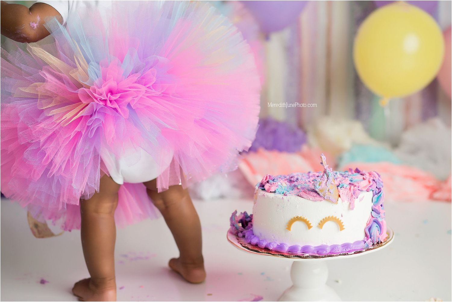 Pastel pink and purple cake smash birthday photo session with tutu, balloons, and decorated cake on white backdrop.