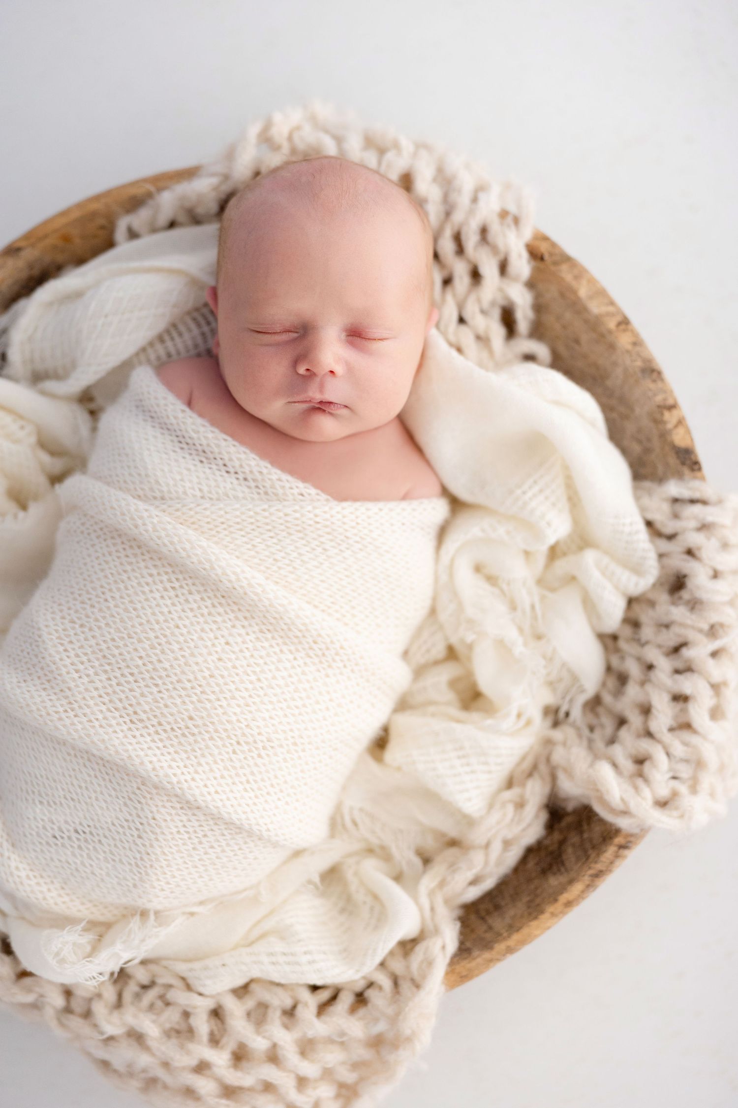 Sleeping newborn baby wrapped in cream knit blanket inside a wooden bowl on white background.