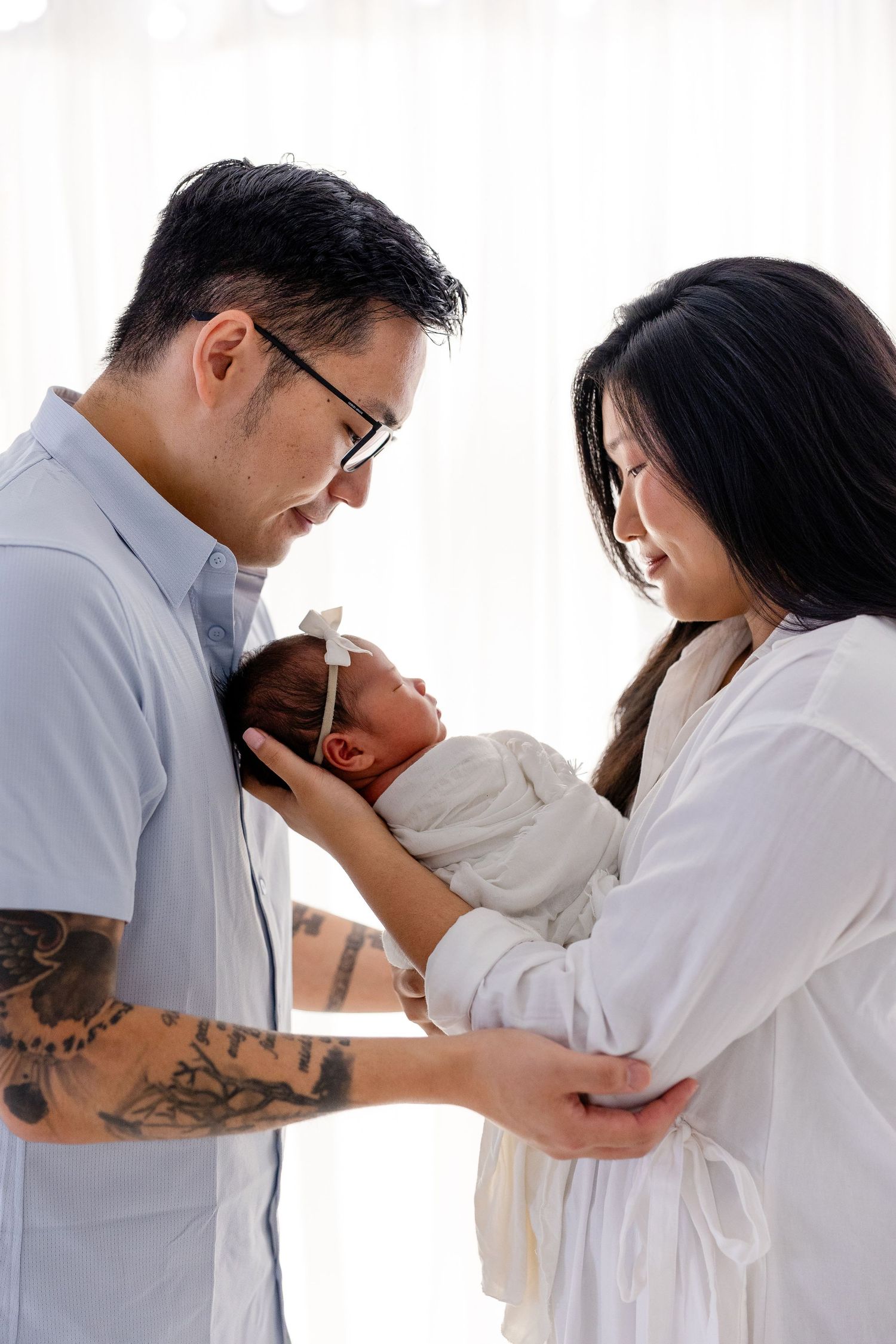 Asian couple lovingly gazing at their newborn baby wrapped in white, father with tattoos wearing glasses, indoor setting.