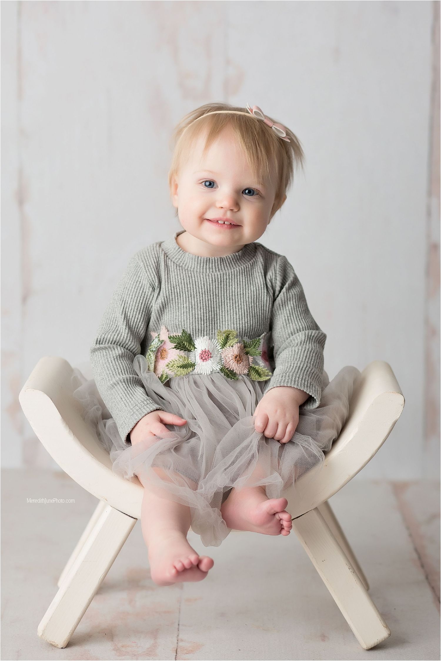 Sweet toddler in gray sweater with floral detail and tulle skirt sits on white wooden chair against light backdrop.