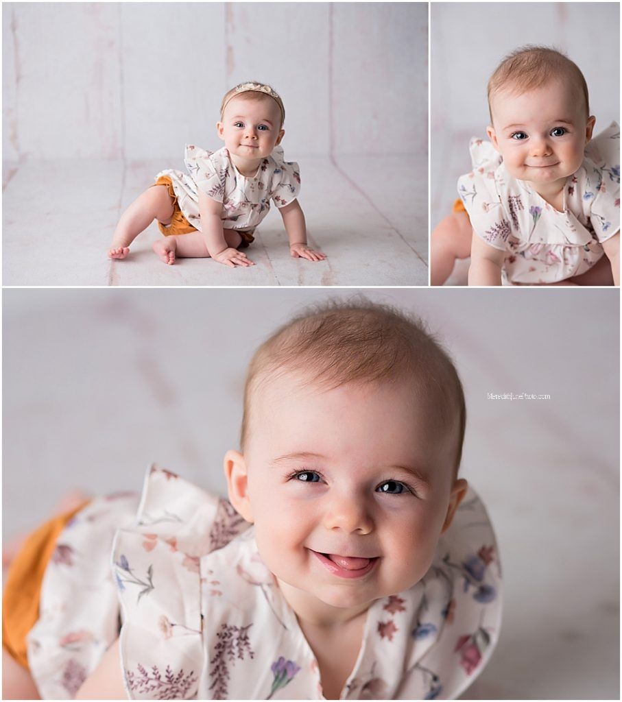 Baby in white floral dress crawls and poses on light gray backdrop during professional photo session.