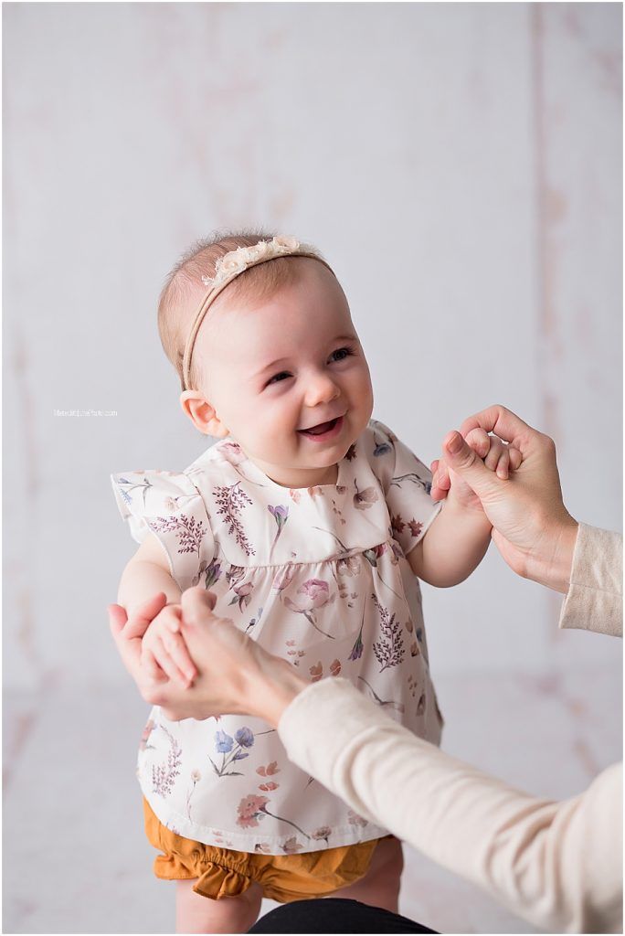 A smiling baby wearing a floral print top and mustard pants reaches up while being held by someone in a cream sweater.