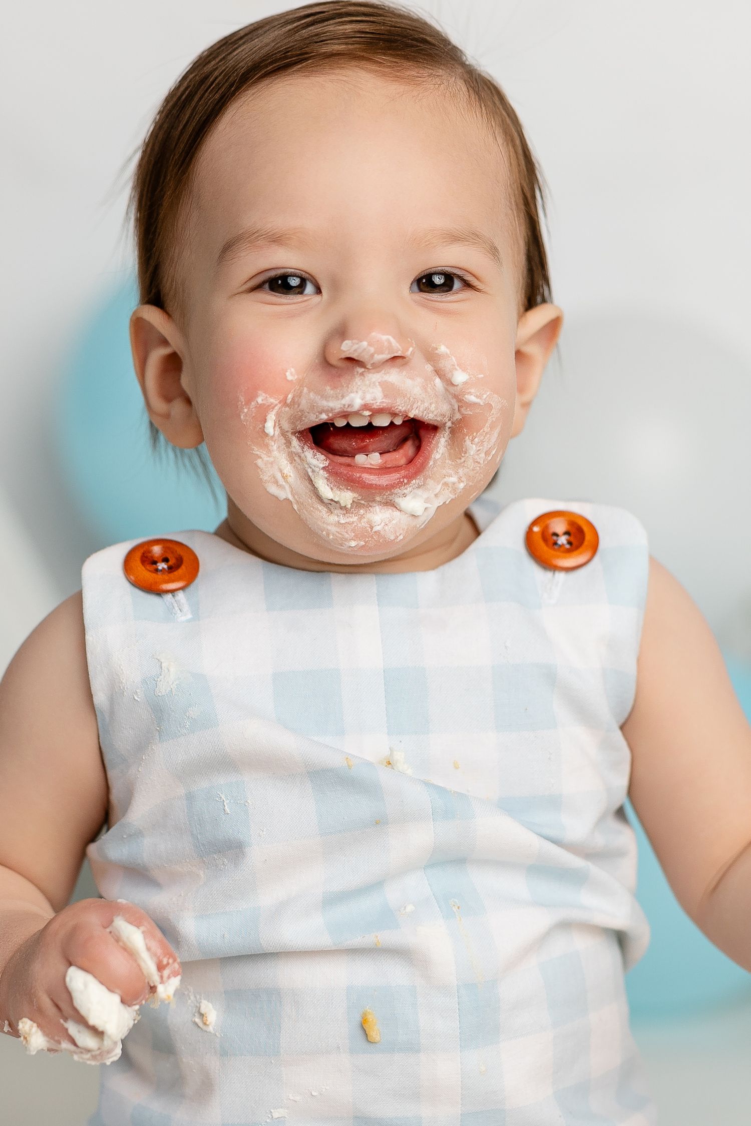 Joyful baby wearing blue and white outfit with orange buttons has messy face covered in white cream or frosting.