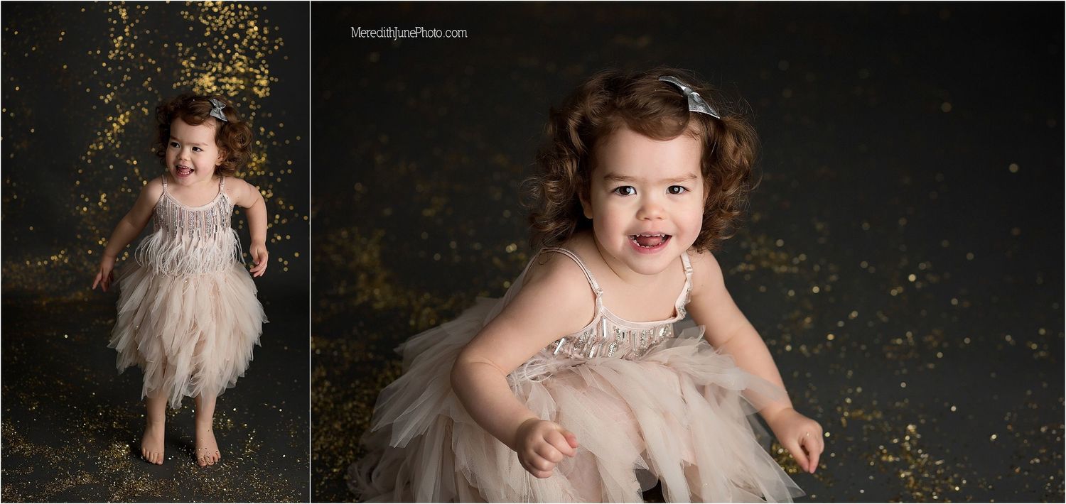 A toddler wearing a sparkly beige tulle dress dances joyfully against a dark backdrop with golden glitter effects.