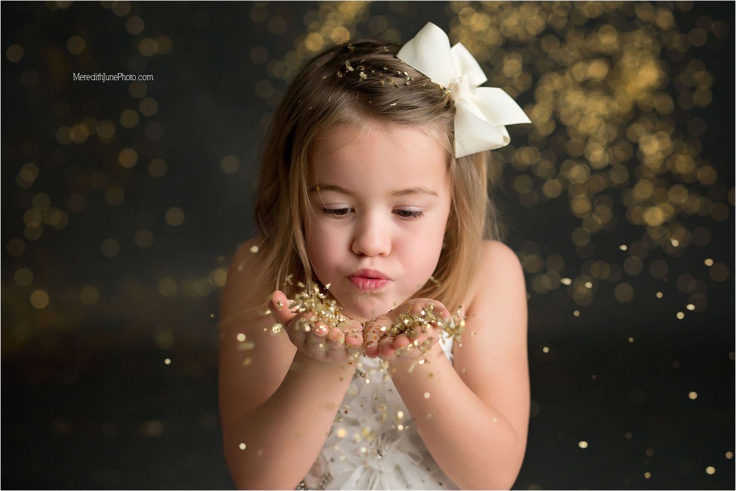 A young child in a white dress blows glitter from their hands against a dark sparkly background.