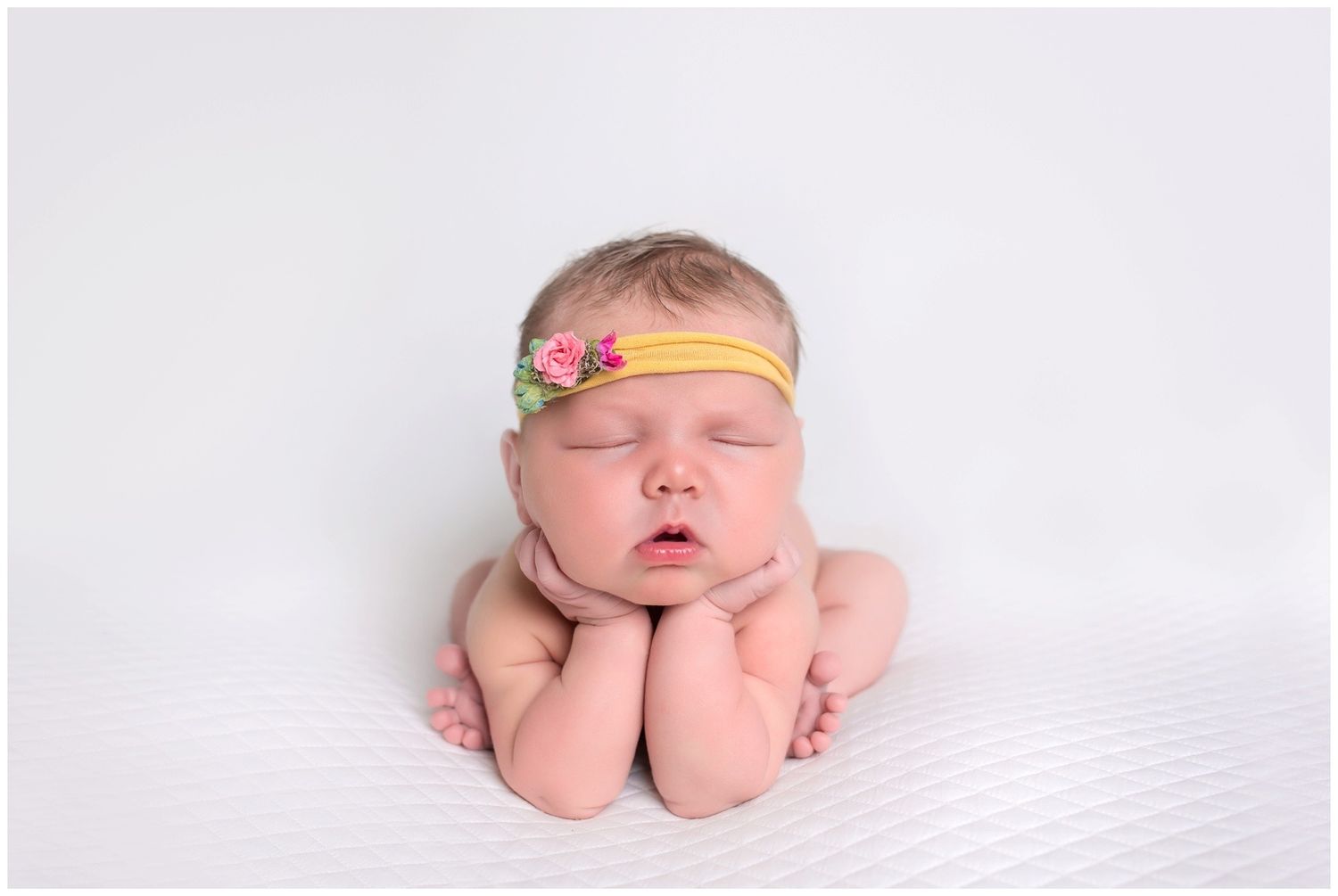 A sleeping newborn baby wearing a delicate floral headband rests peacefully on soft white blankets.