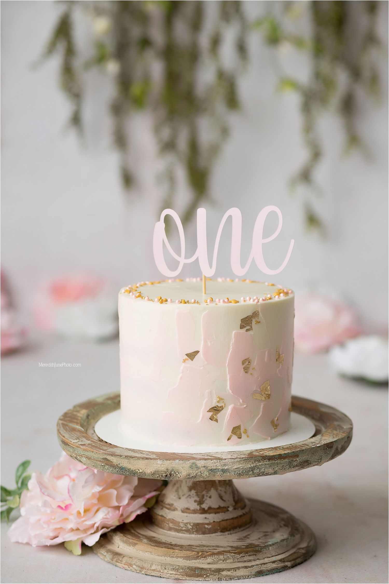 Elegant pink buttercream cake with gold accents displayed on a rustic wooden cake stand with soft floral decorations.