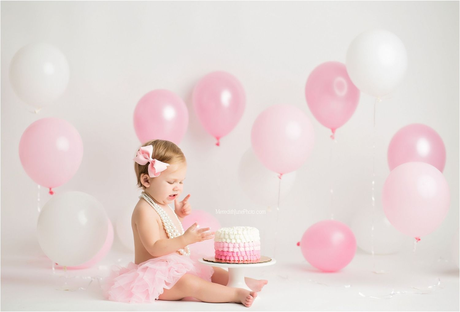 Baby in pink tutu dress sits among floating pink balloons during first birthday cake smash photo session.