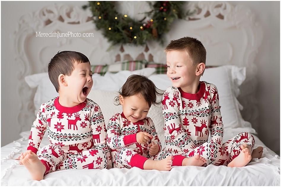 Three children in matching red and white Nordic print Christmas pajamas laugh together on a bed with holiday decor behind them.