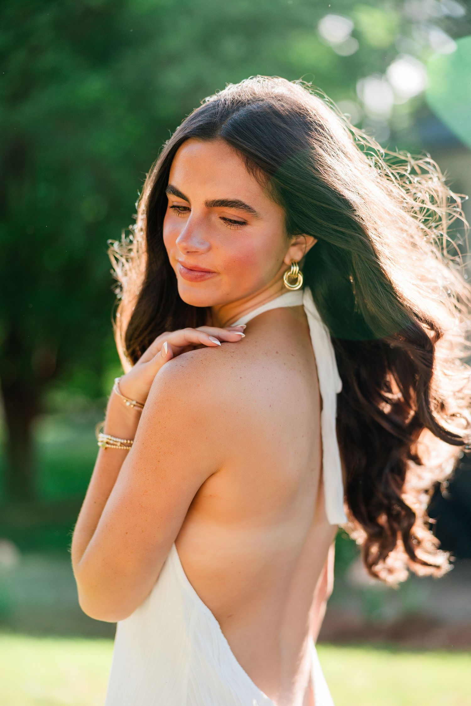 Young woman in backless white dress with gold earrings and bracelet, posing outdoors in sunlit green garden.