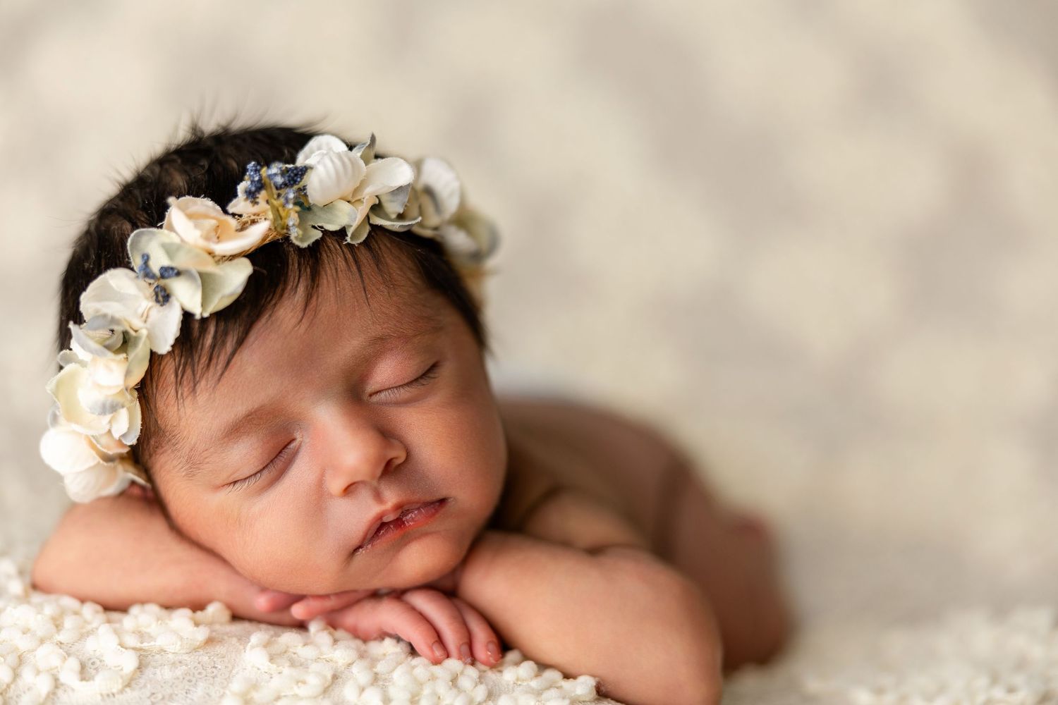 Sleeping newborn baby wearing delicate white floral crown rests peacefully on cream textured blanket.